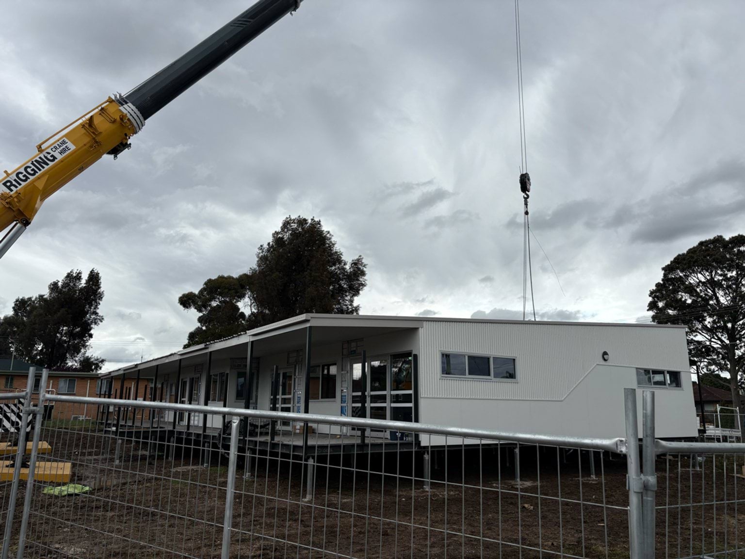 A modular building being dropped into place on the St Albans West Kindergarten site by a crane. There is a metal protective fence in front of it. 