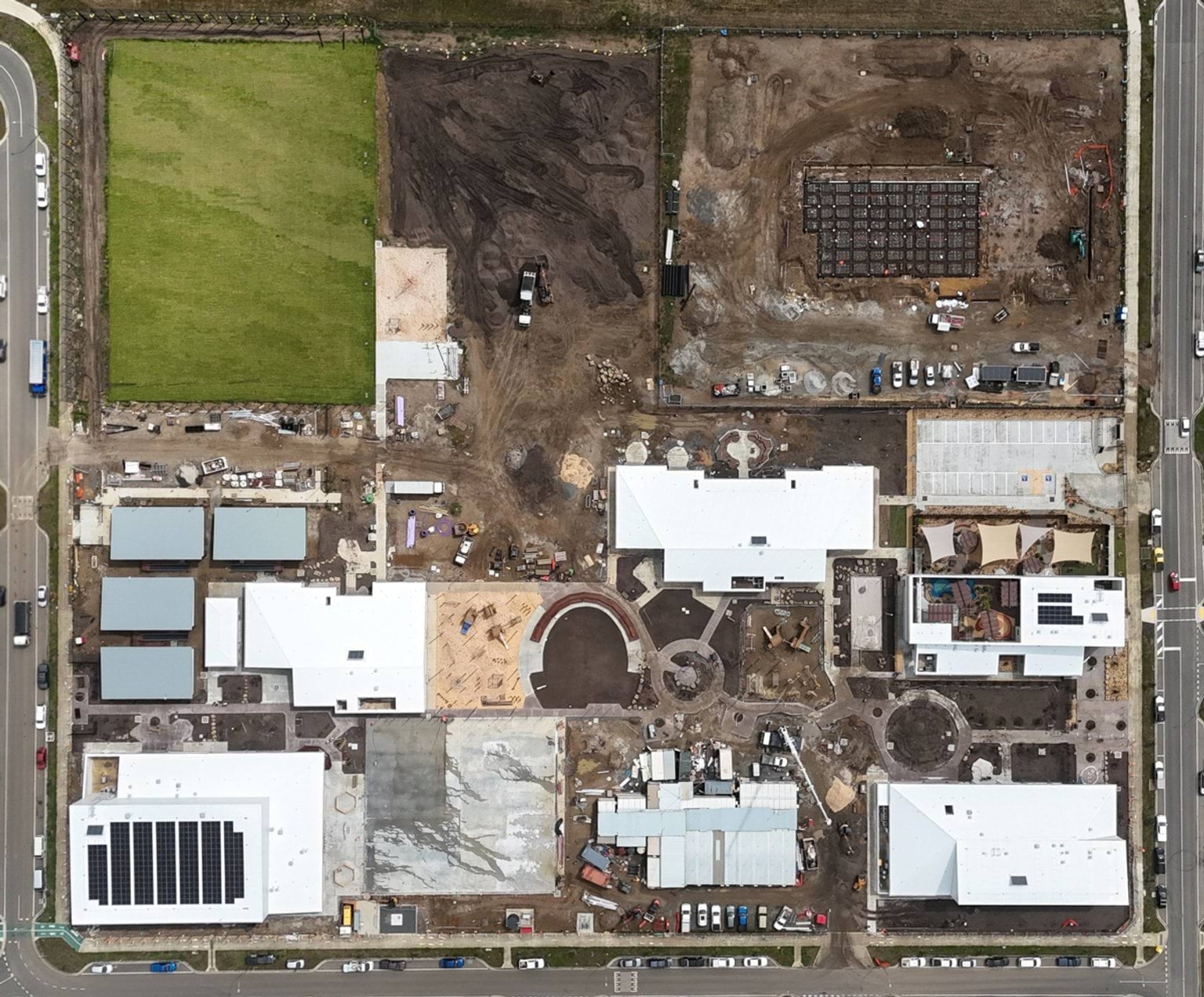 An aerial photo of the Kulap Primary School construction site. There is a green grass rectangle field in the top left, with a dirt patch to its right. There are various buildings in the area below, mostly looking finished from the outside. There is also a concrete car park. Bordering the construction site are roads.
