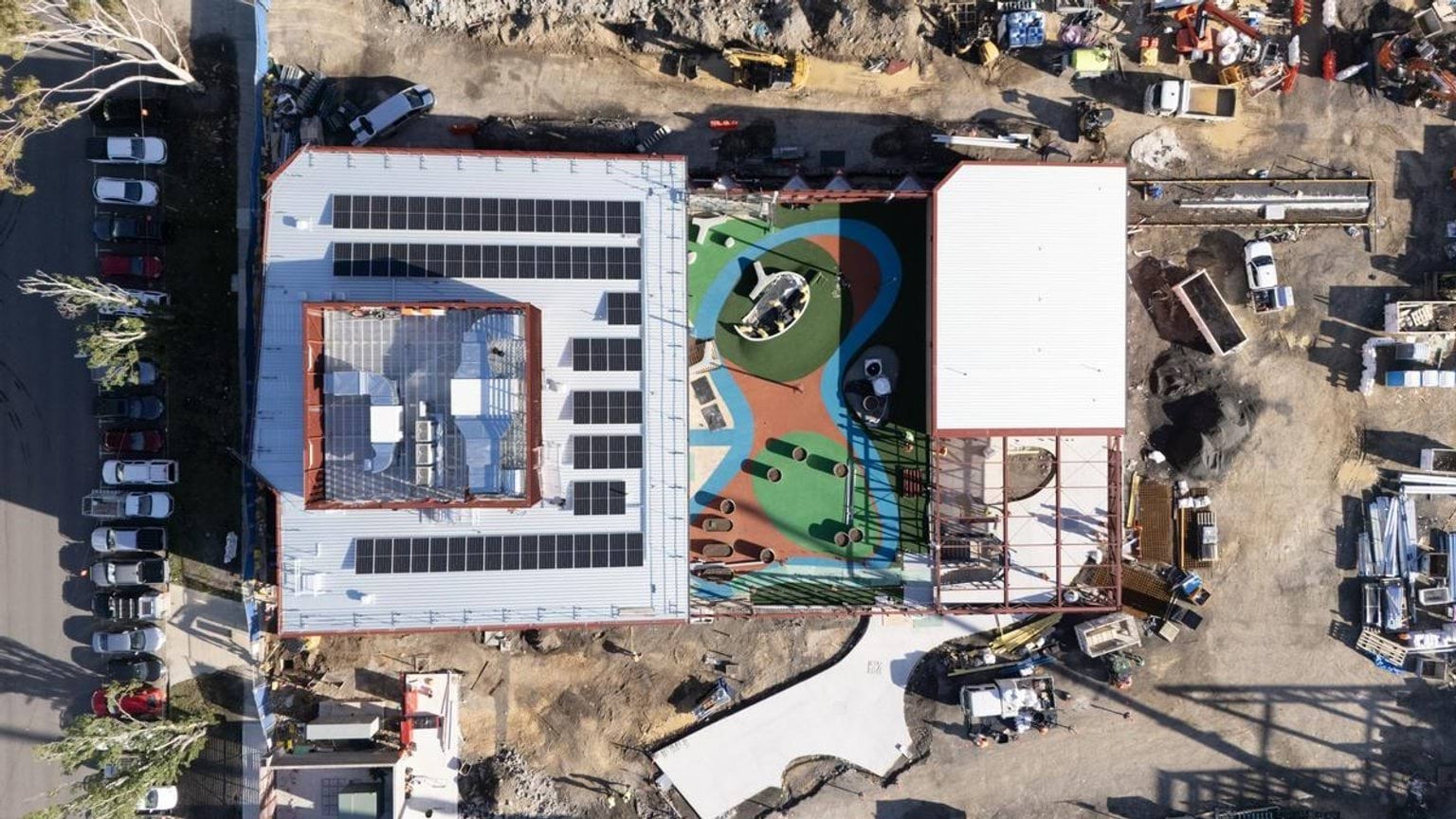 An aerial photo of the Narrarrang Primary School Kindergarten construction site. The buildings looks almost finished. There are solar panels on one roof, and red/green/blue coloured ground between the 2 buildings, with a concrete pathway leading out of one of them. The rest of the site is filled with vehicles, machinery and construction materials on dirt. 