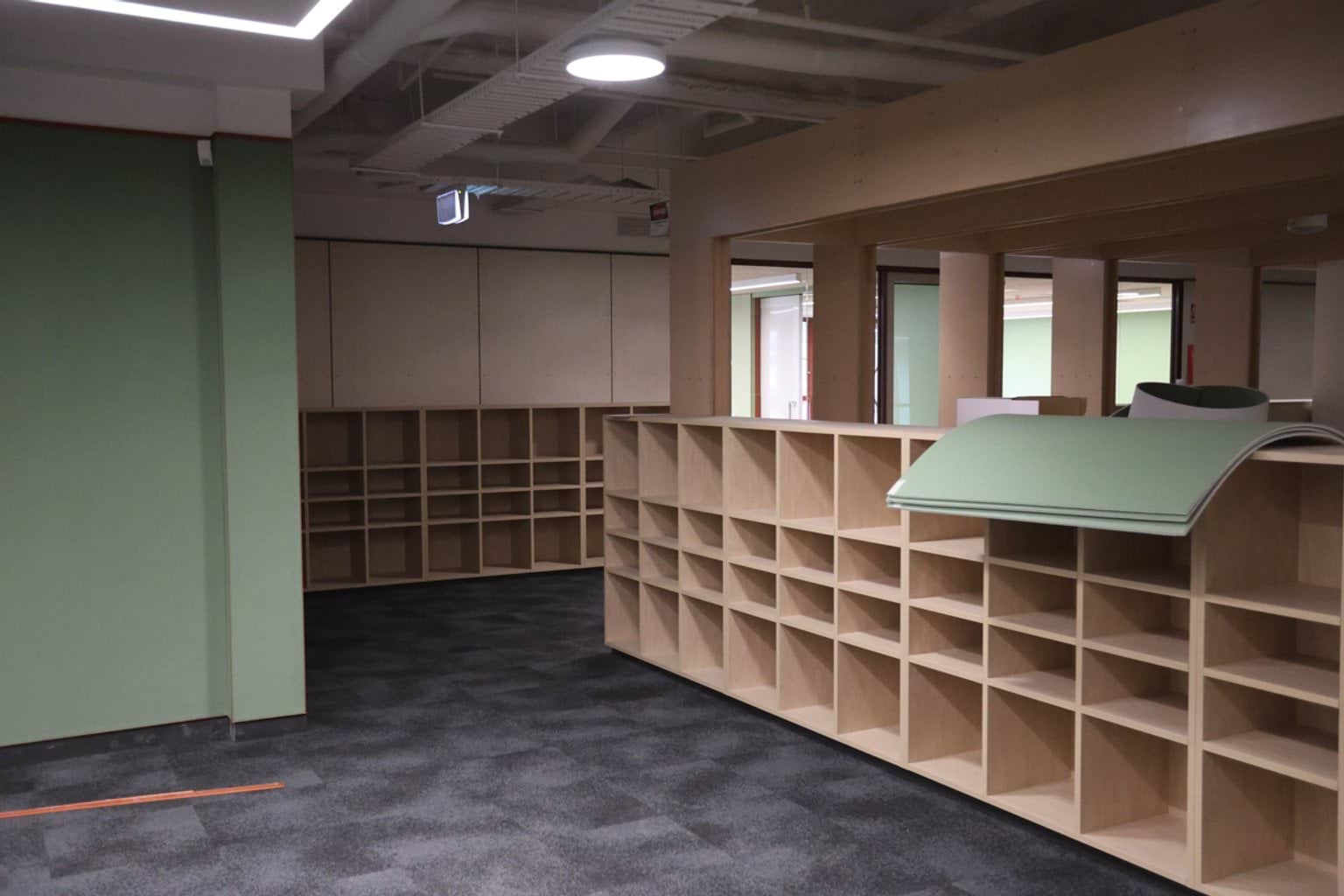 Inside school room. There are wood shelves on the walls and overhead lights. Some green panels are on the shelves.
