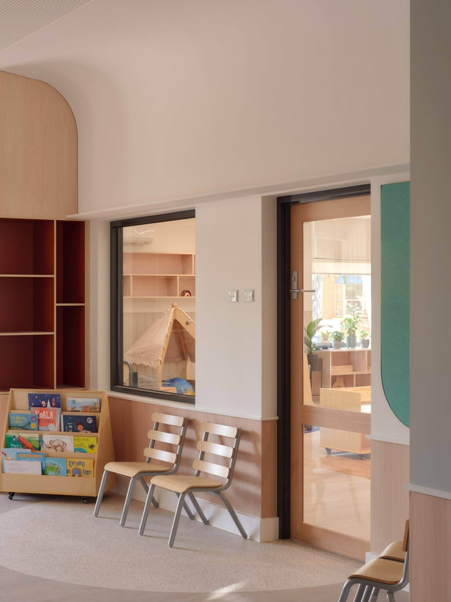 The interior of the kindergarten, showing a light-filled hallway with timber wall panels and two chairs against the wall. A book display with colourful children’s books is on the left, and through the window there is a small wooden playhouse.
