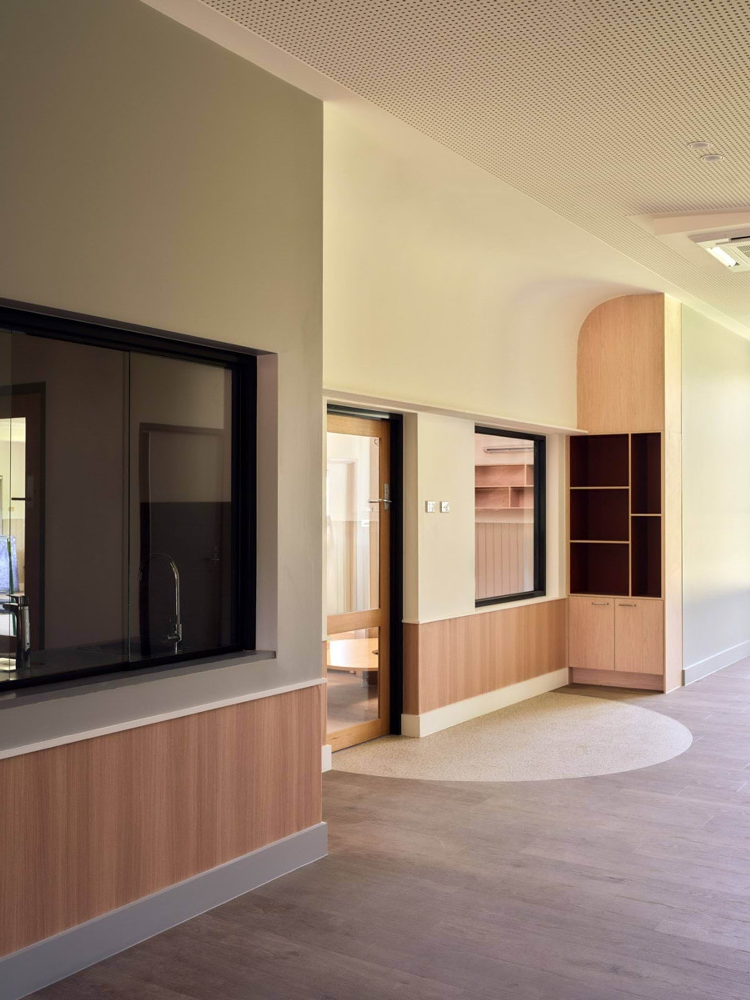 A hallway inside the kindergarten with timber wall panels and built-in shelving. There are large windows looking into adjoining rooms and a wooden door with glass panels.