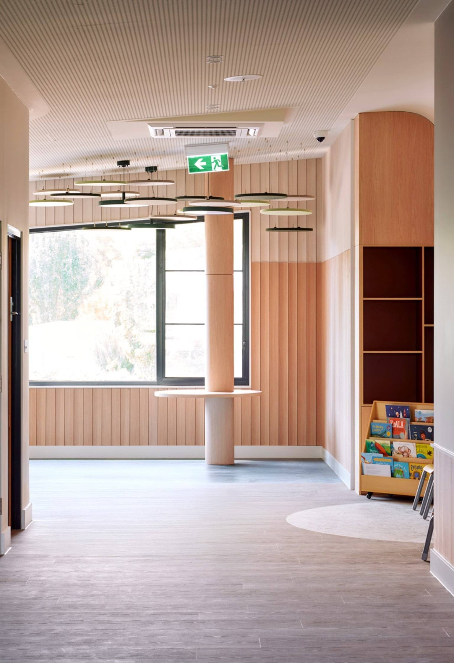 A bright hallway inside the kindergarten with timber wall panels and a large window. Circular light fittings hang from the ceiling and a book display sits in the corner.