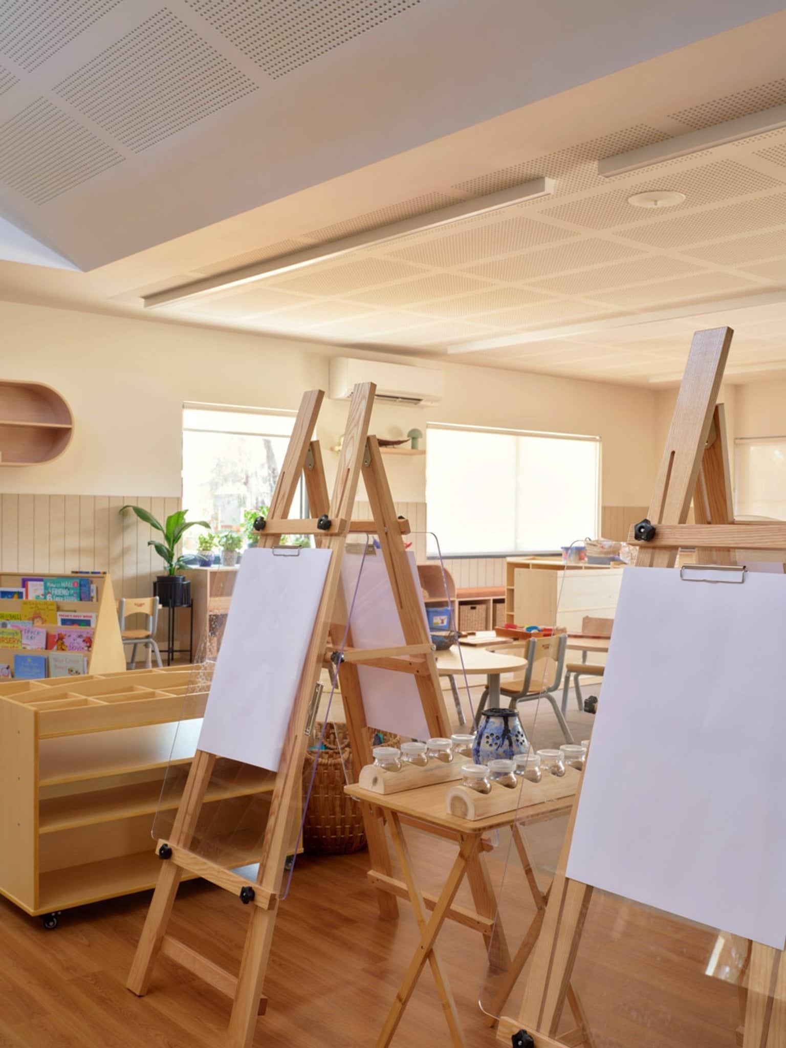 A close-up of easels in a kindergarten classroom, with blank sheets of paper ready for painting. There are small tables with jars and brushes, and shelves in the background holding books and art supplies.