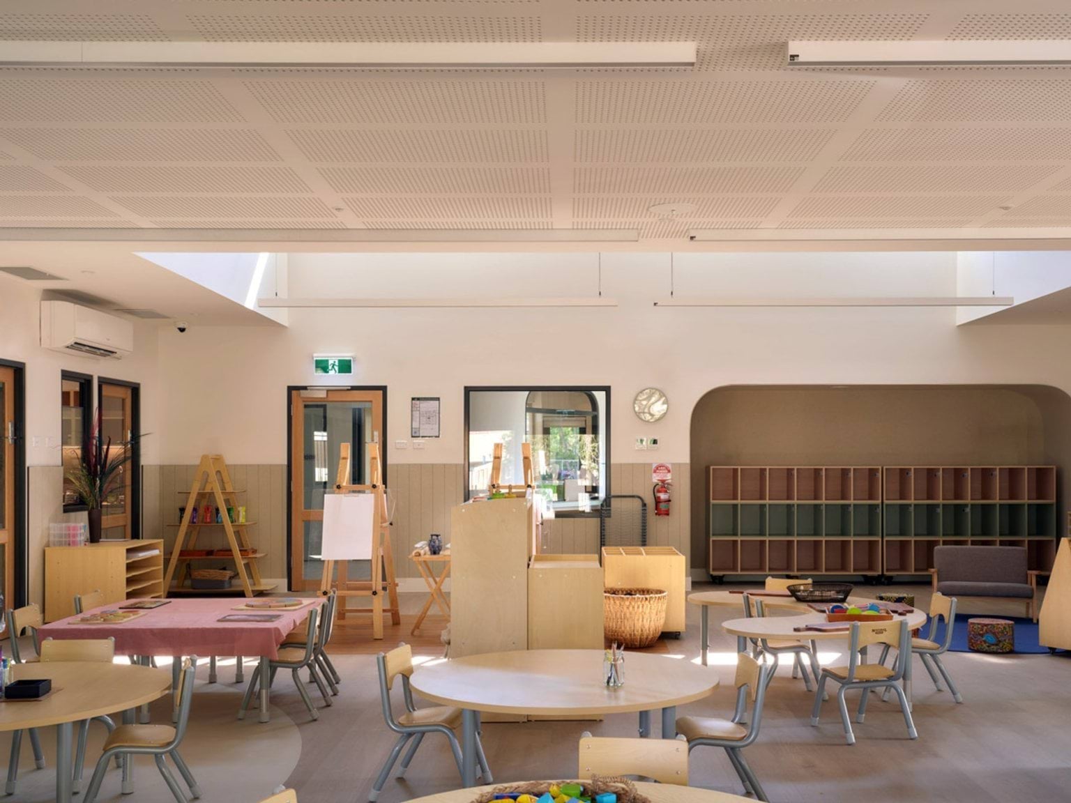 A kindergarten classroom with round tables and small chairs arranged in the centre. There are easels, shelves, and storage cubbies along the walls, and natural light coming through high windows.