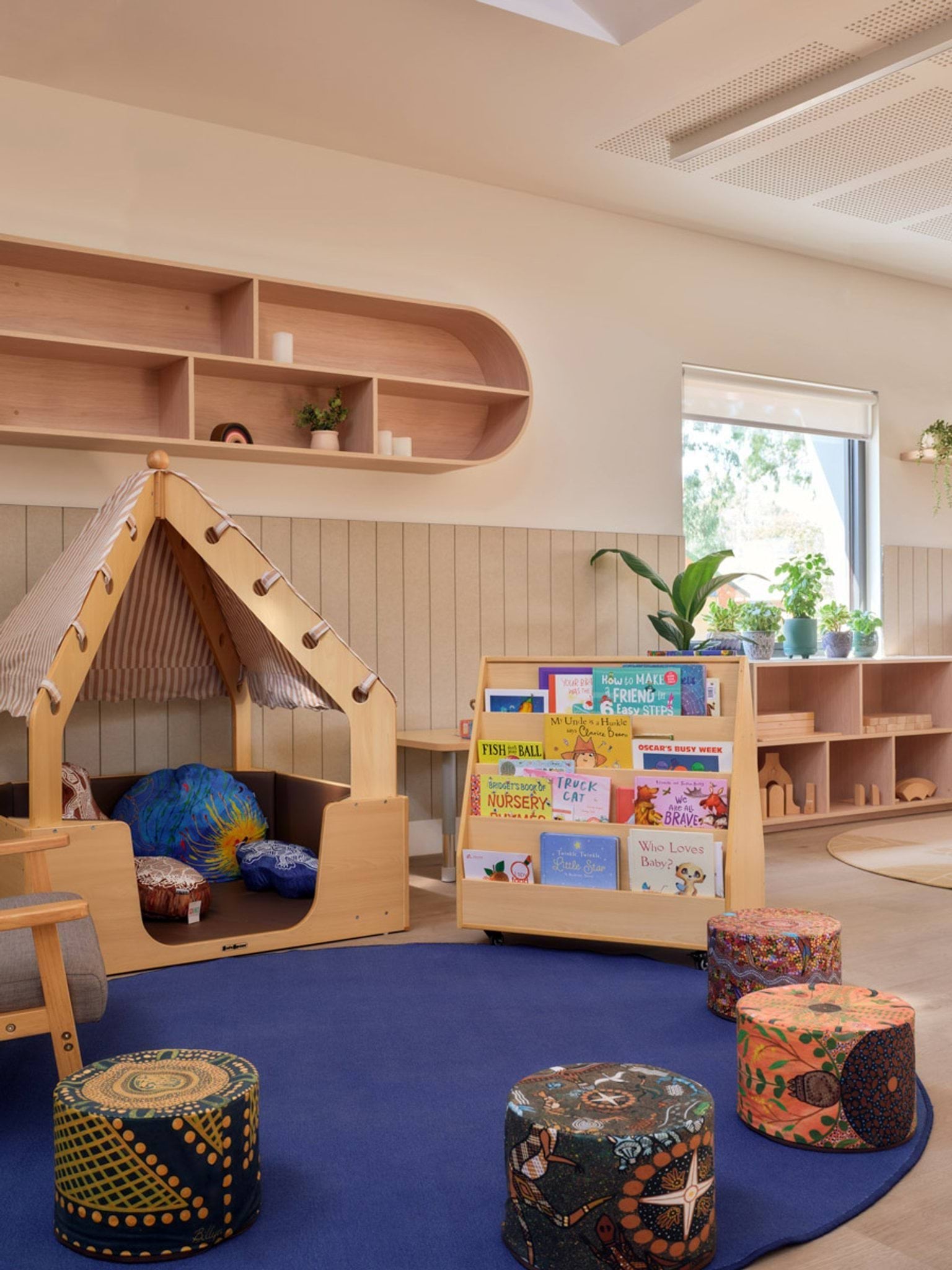 A reading corner inside the kindergarten with a small wooden cubby house, a blue rug, and round stools with colourful patterns. A book display holds picture books and there are plants on shelves near the window.