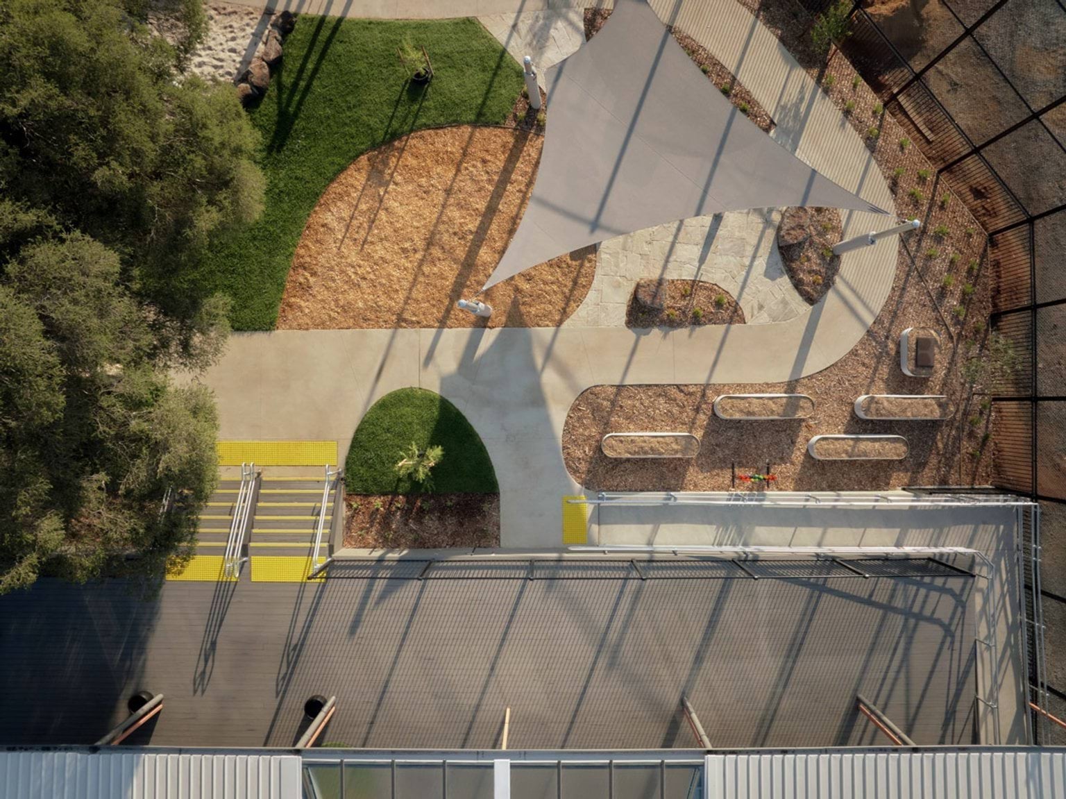 An aerial view of the kindergarten outdoor play area, with curved concrete paths, garden beds, and a large shade sail. There are timber benches and a mix of grass and mulch surfaces.