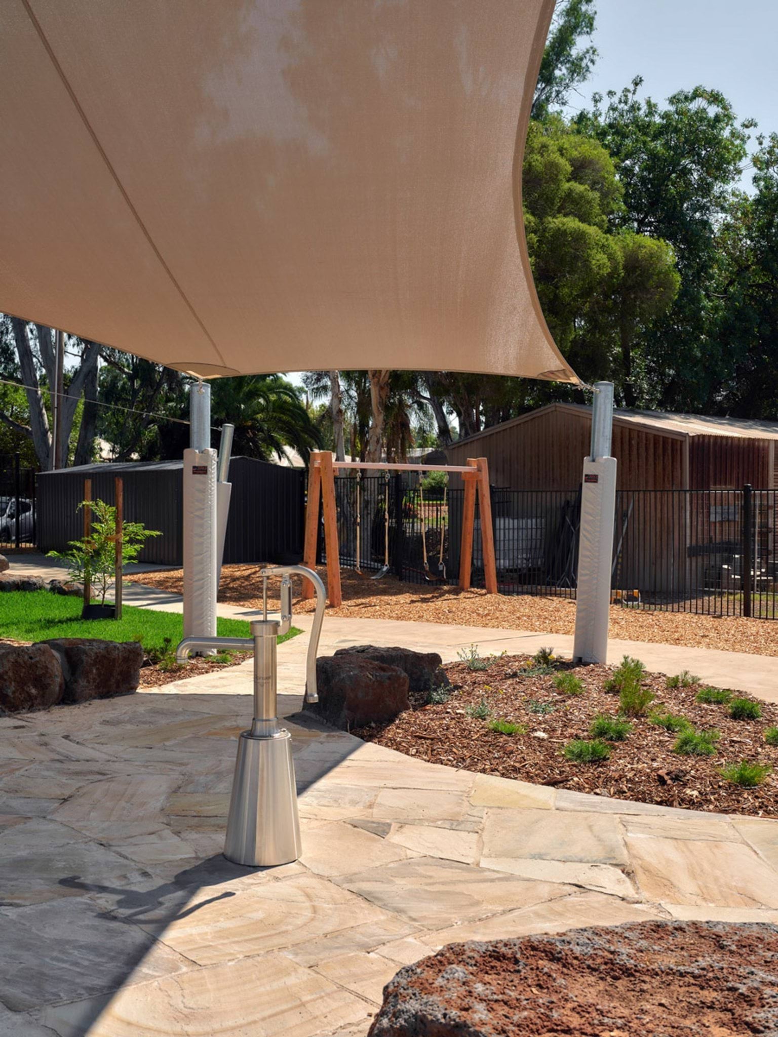 The outdoor play area of the kindergarten, showing a stone-paved surface with a stainless steel water pump. A large shade sail stretches overhead and there are garden beds and rocks nearby.