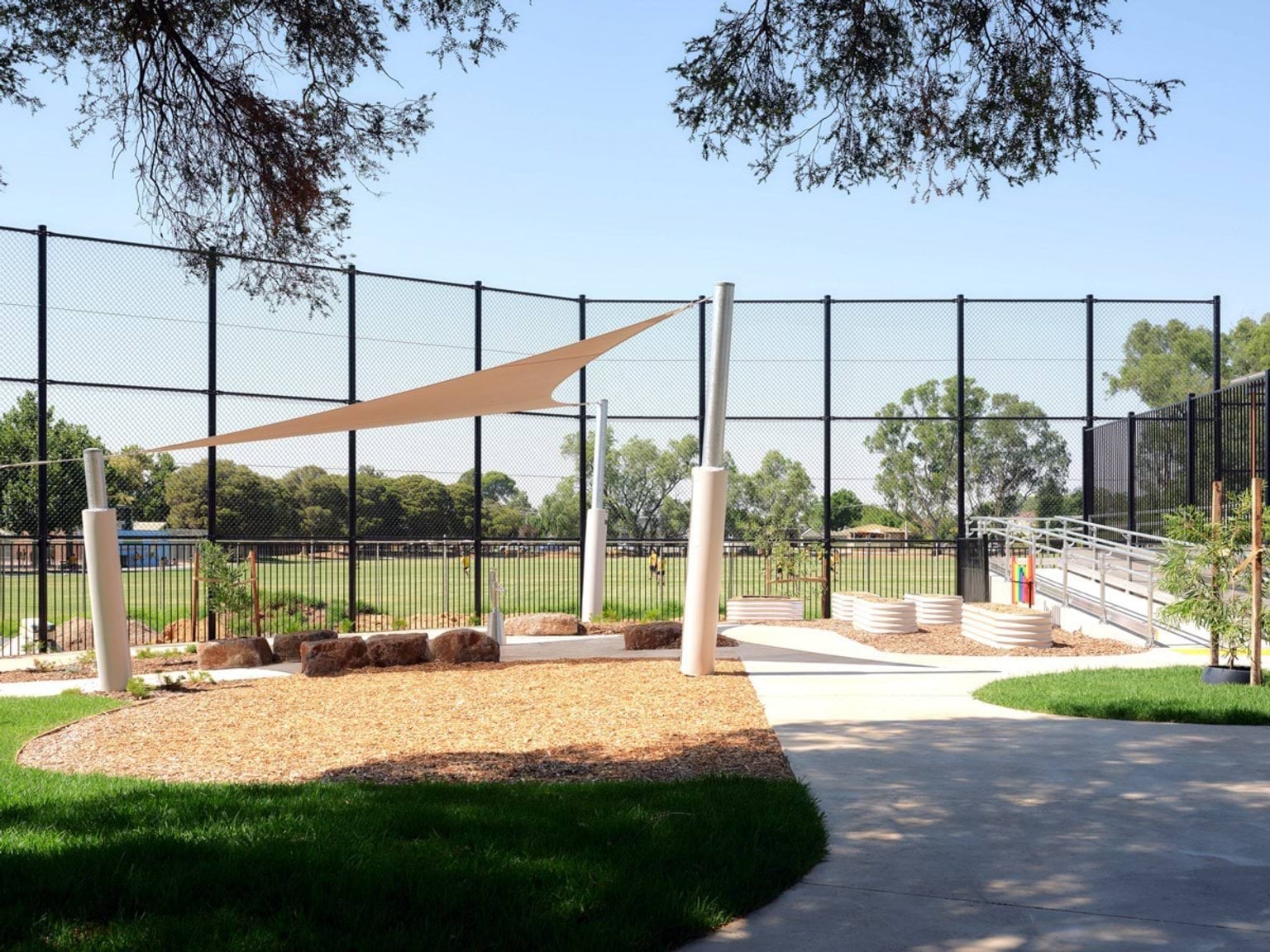 The kindergarten outdoor play area with a shade sail, garden beds, and a curved concrete path. A black mesh fence encloses the space and trees are visible beyond.