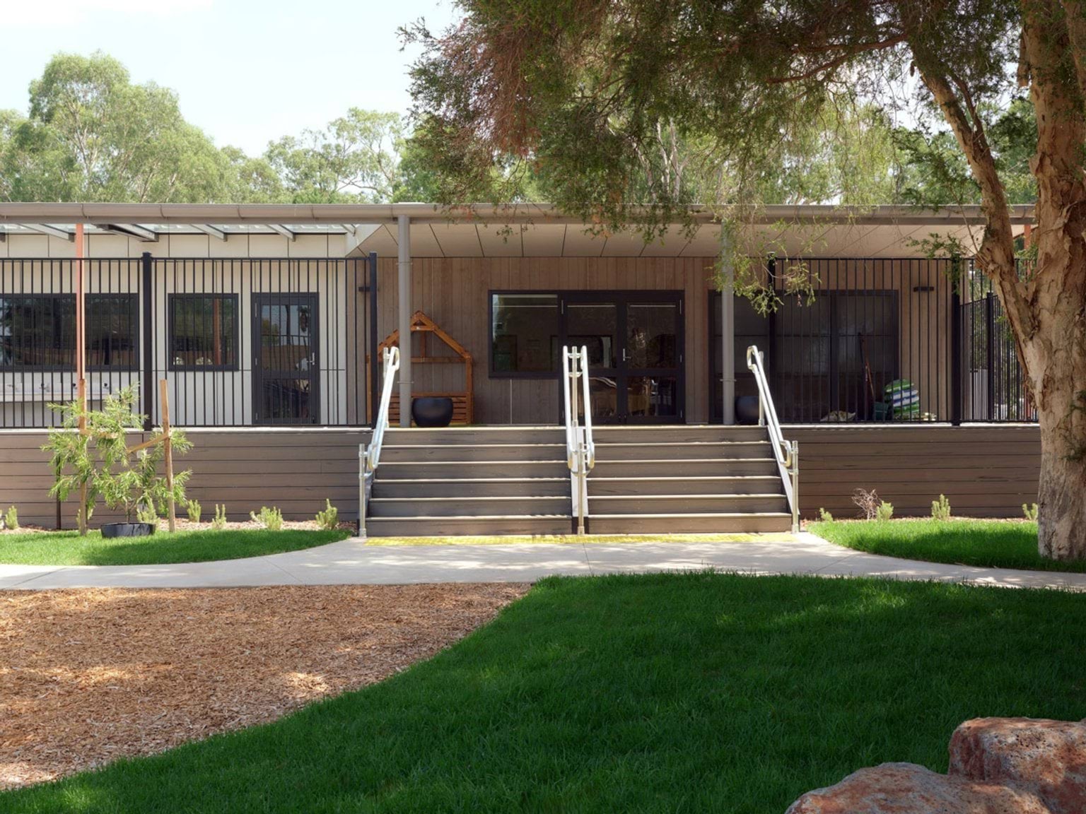 The exterior of the kindergarten viewed from the garden, with timber walls and a covered veranda. Wide steps lead up to the entrance and there is a large tree in the foreground.