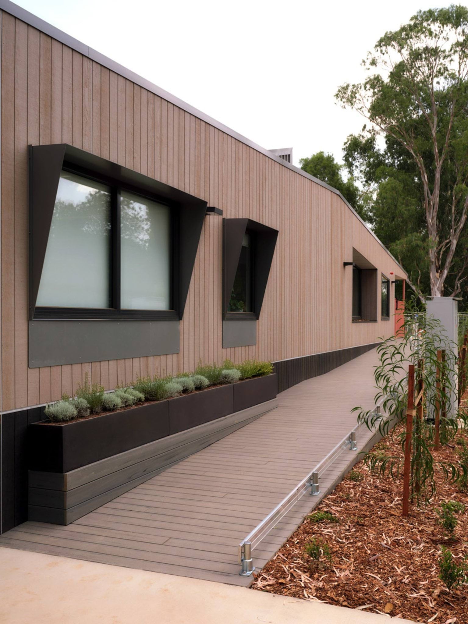 The exterior of the kindergarten, which has light timber cladding and angular black window shades. A timber walkway runs along the building with planter boxes and young trees planted in mulch beds.