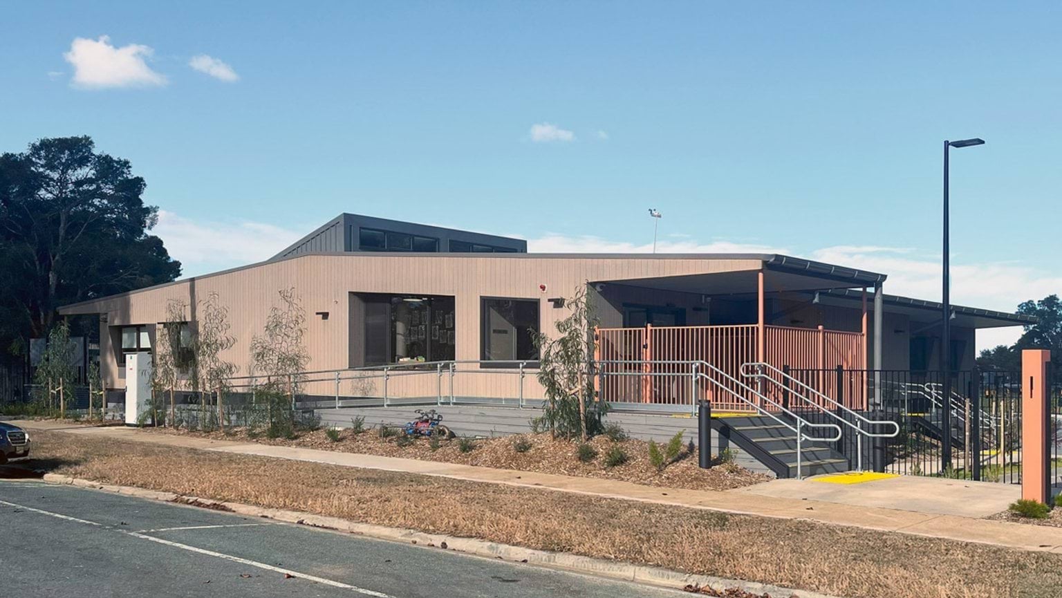 The exterior of the kindergarten, which has light timber walls and a covered veranda with orange railings. There is a ramp and stairs leading up from the footpath, and young plants in garden beds.