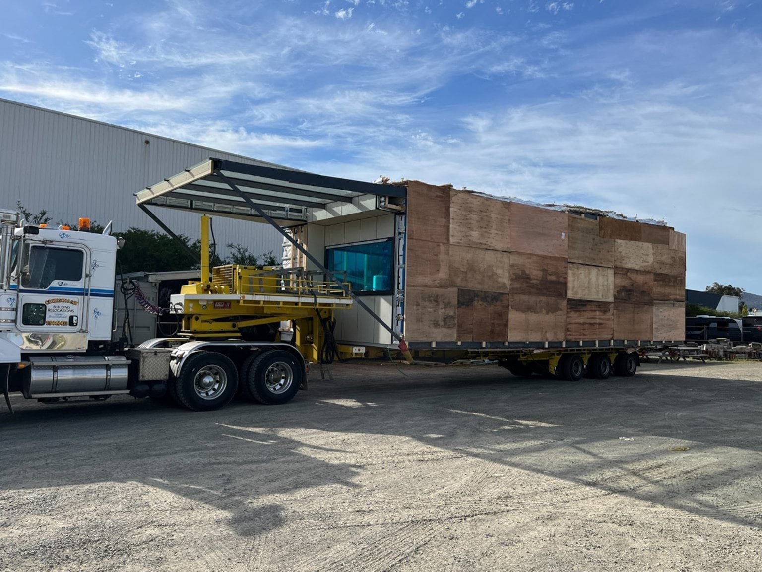 A prefabricated kindergarten module loaded onto a truck for transport. The module is wrapped in plywood panels and secured with straps, and the truck is parked on a gravel surface.