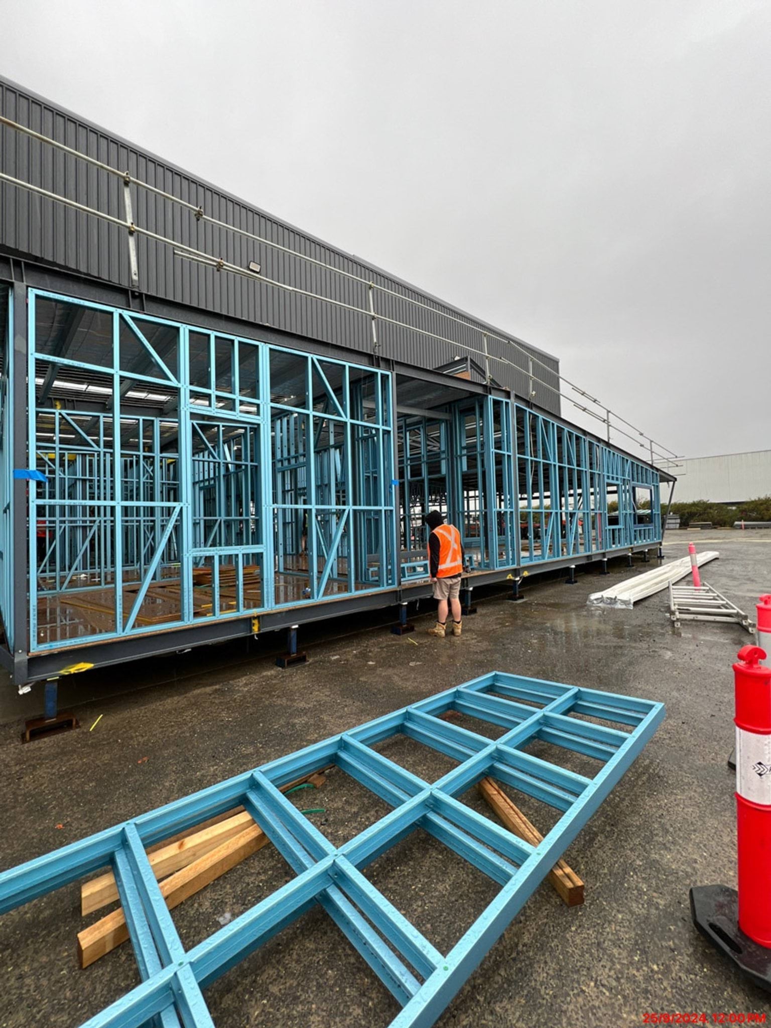 A kindergarten module under construction, showing blue steel framing and a worker in a high-visibility vest. Timber pieces and tools are scattered on the ground nearby.