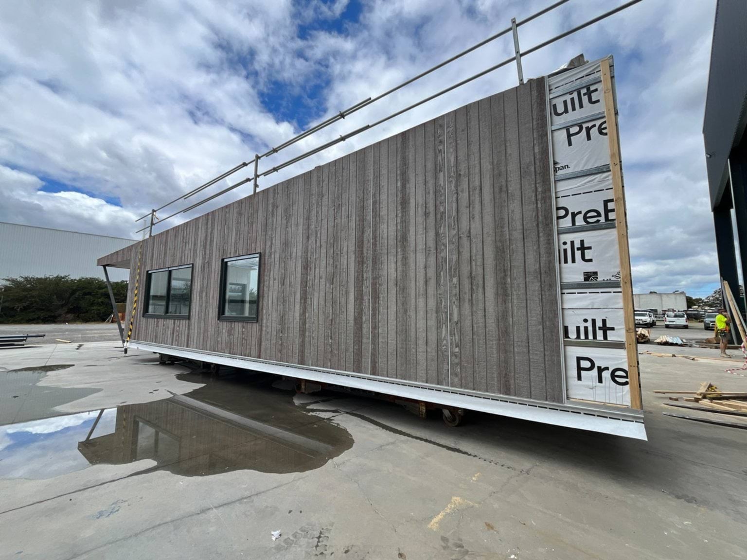 A prefabricated kindergarten module under construction, with timber cladding on one side and insulation wrap visible on the other. The module is sitting on a concrete surface with scaffolding above.