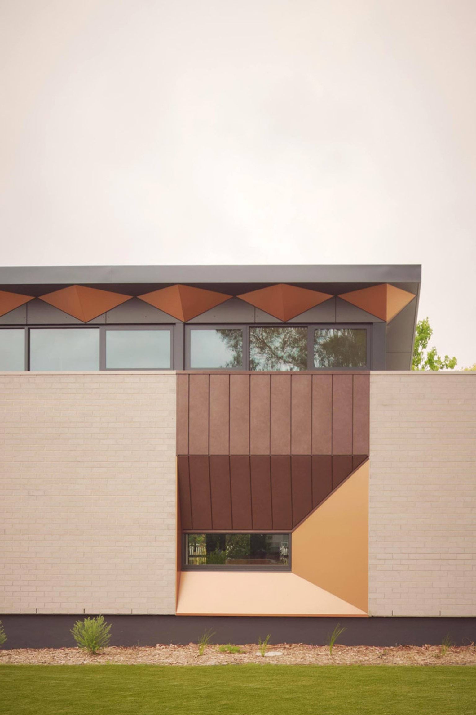 A close-up of the kindergarten exterior showing a feature window with angled copper panels and cream brick walls. There are small plants in a mulched garden bed below.