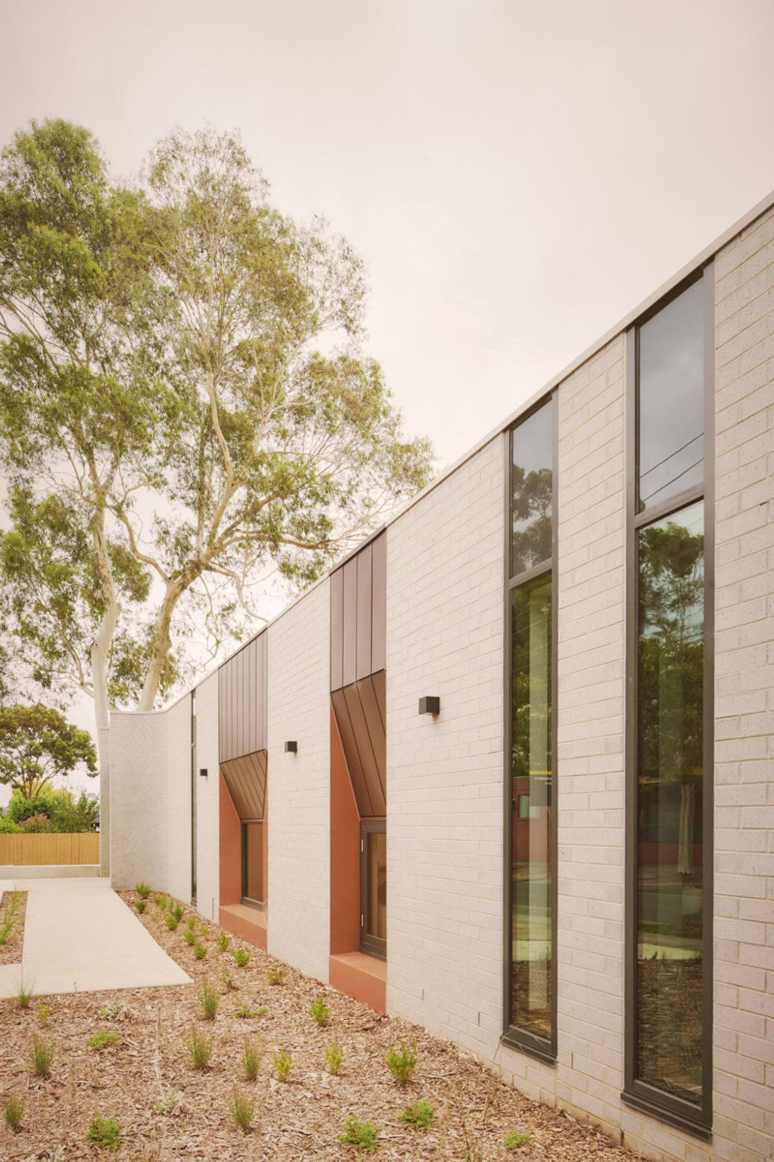 The exterior of the kindergarten with tall narrow windows and angled copper panels set into cream brick walls. A concrete path runs alongside a garden bed with small plants.