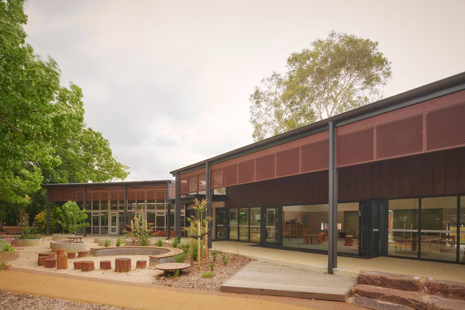 The exterior of the kindergarten with rust-coloured screens and dark cladding. A landscaped outdoor play area has timber log seats arranged in a circle and garden beds with young plants.