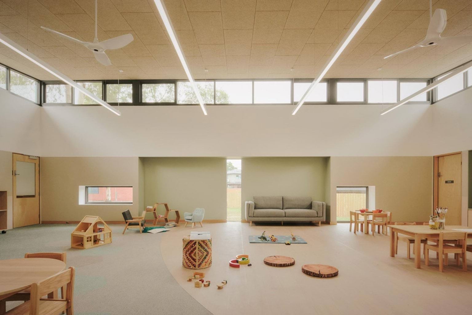 A kindergarten classroom with high windows and a pitched ceiling. There are small timber tables and chairs, a grey couch, and play areas with wooden toys and cushions on the floor.