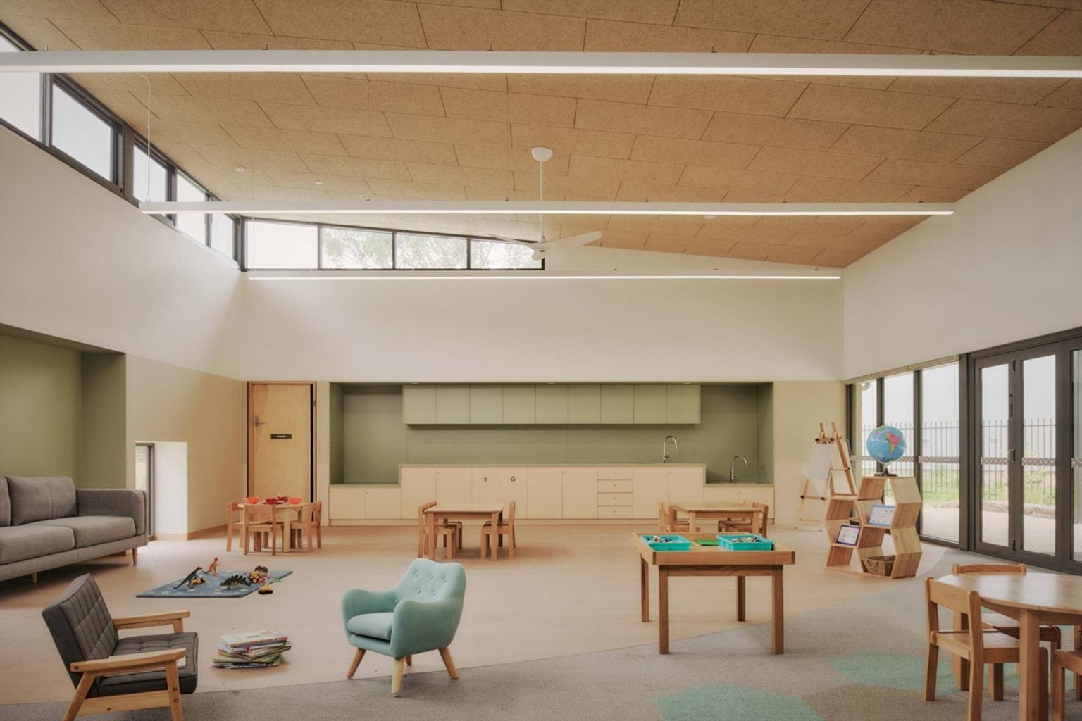 A kindergarten classroom with timber furniture, a grey couch, and a play table. Built-in cabinetry runs along the back wall and large windows bring in natural light.