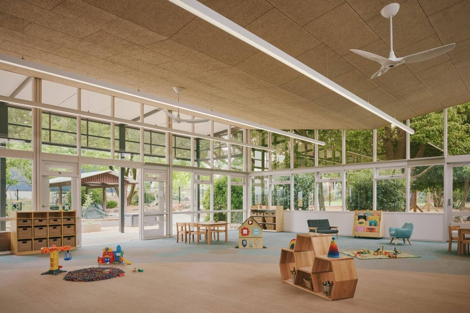 A kindergarten classroom with large windows looking onto a garden. There are timber tables and chairs, play mats, and shelves with toys and books.