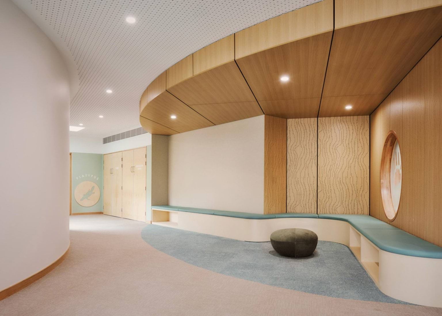 A kindergarten hallway with curved walls and timber ceiling panels. There is teal bench seating along the wall and a round feature window. A door at the end has a platypus illustration.