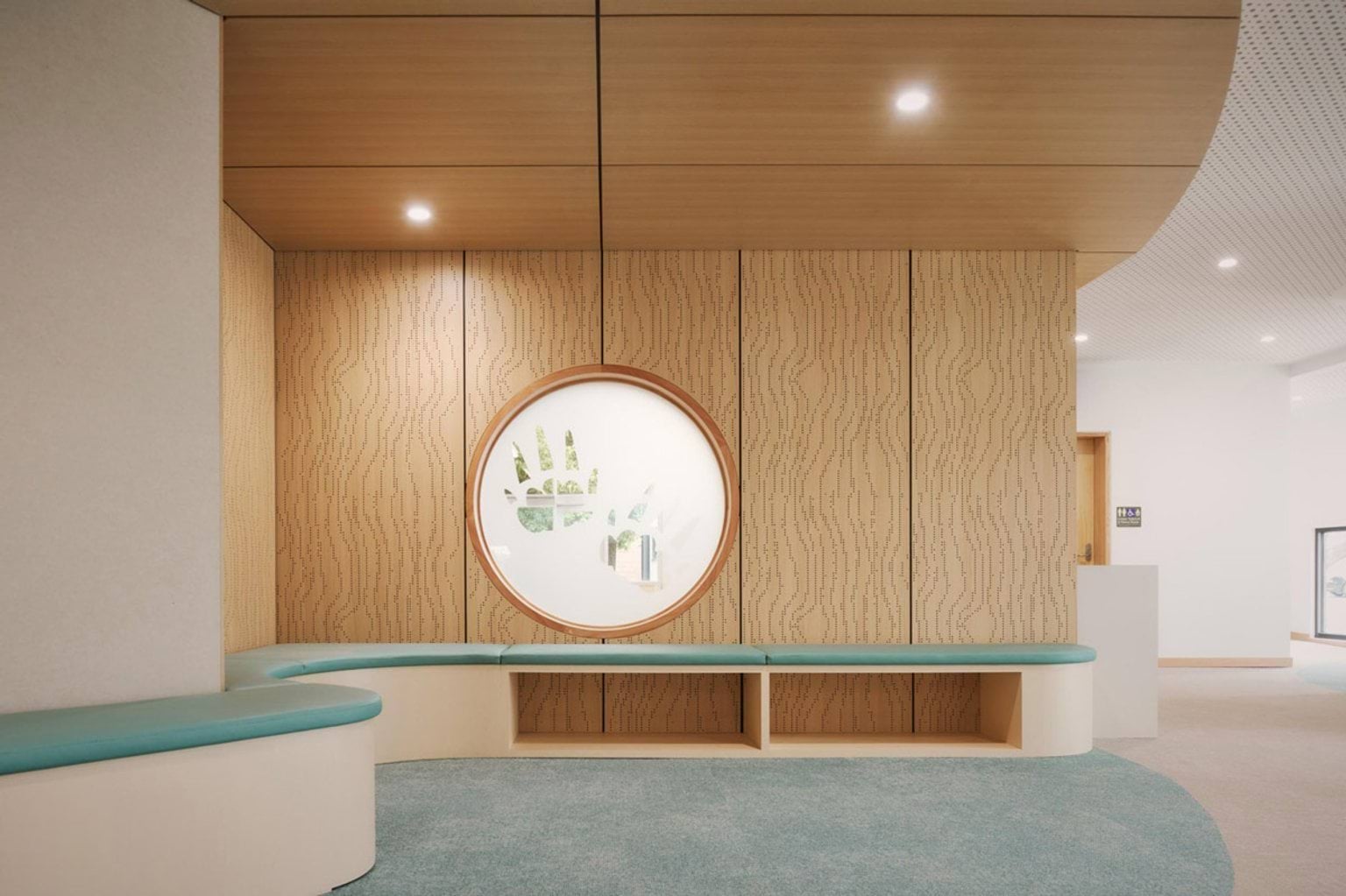 A curved hallway inside the kindergarten with timber wall panels and teal bench seating. A round window with a handprint design is set into the wall.