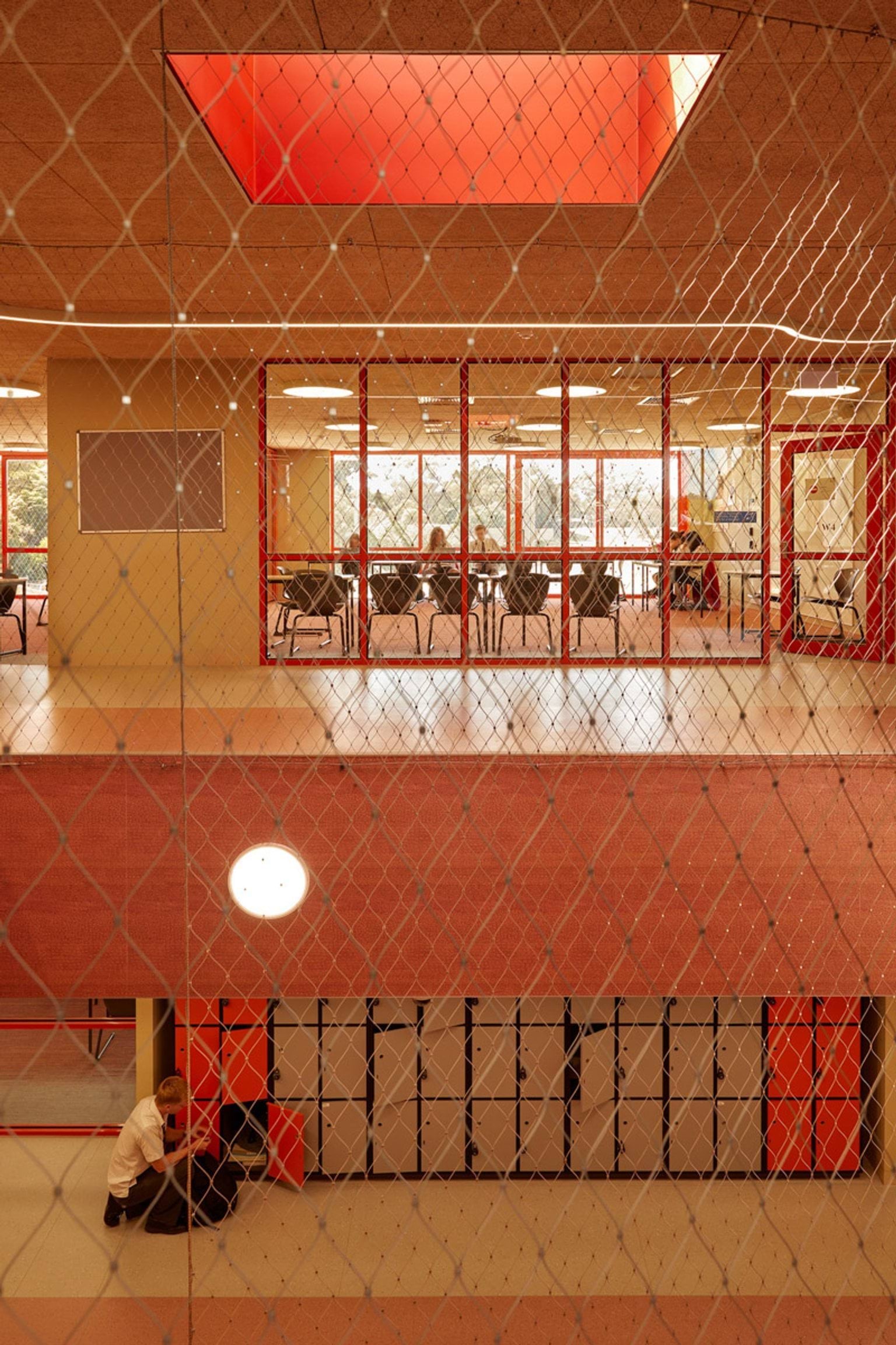 A view through mesh panels into a school interior with red-framed glass walls. Students are seated at desks in a classroom on the upper level, and lockers are visible below.