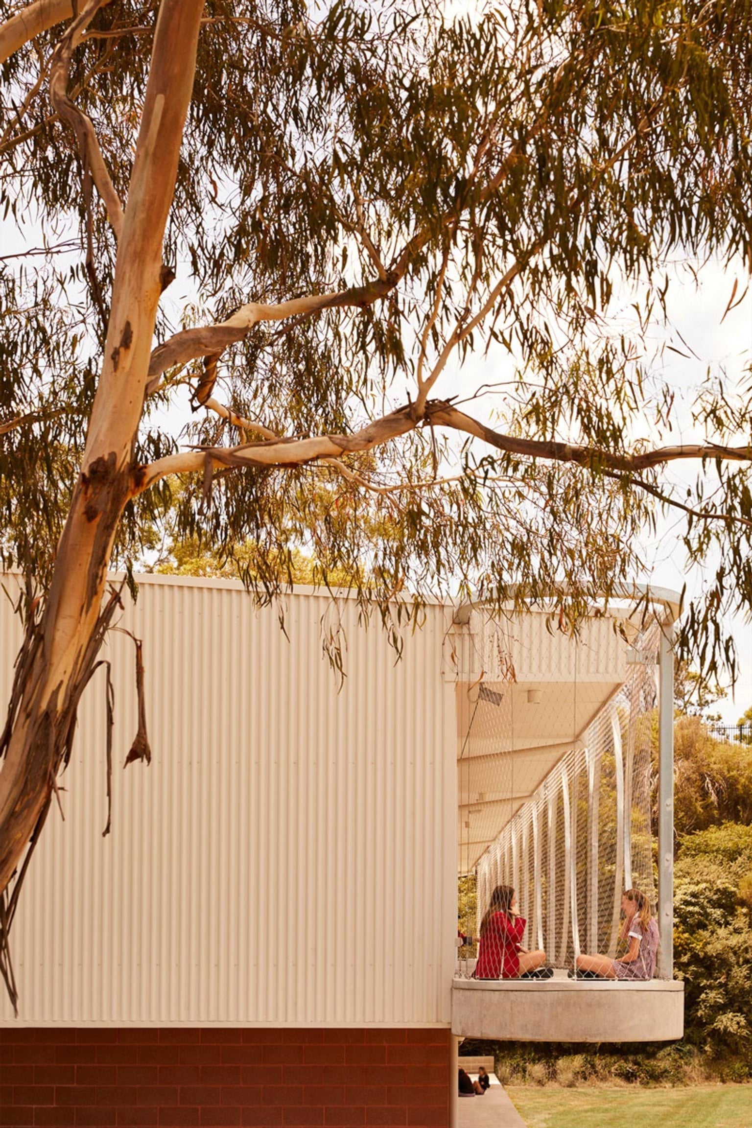 An outdoor balcony with curved metal frames and mesh panels attached to a school building. Two students are seated on the balcony, partially shaded by a tree.