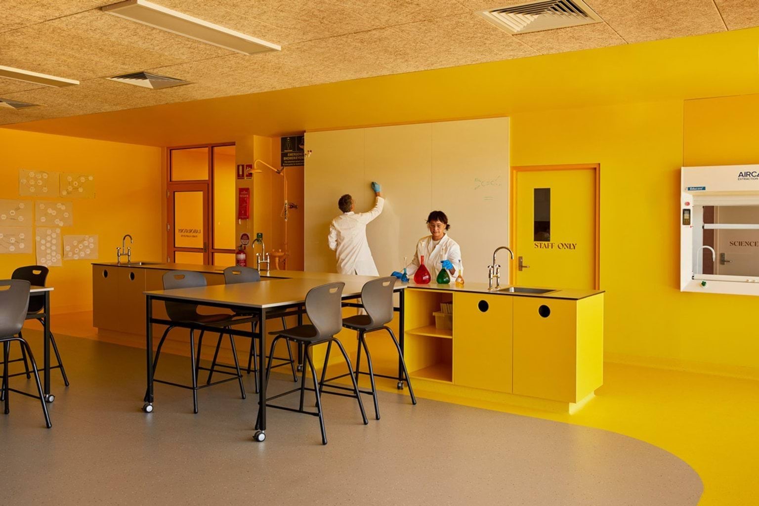 A bright science classroom with yellow walls, black benches, and stools. Two people in lab coats are working at the front bench near a whiteboard.