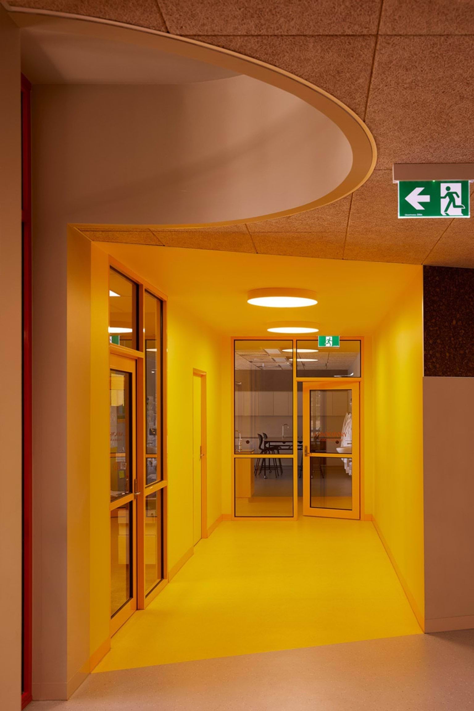 A bright yellow hallway with curved ceiling detail and glass doors leading to a classroom. An exit sign is mounted above the doorway.