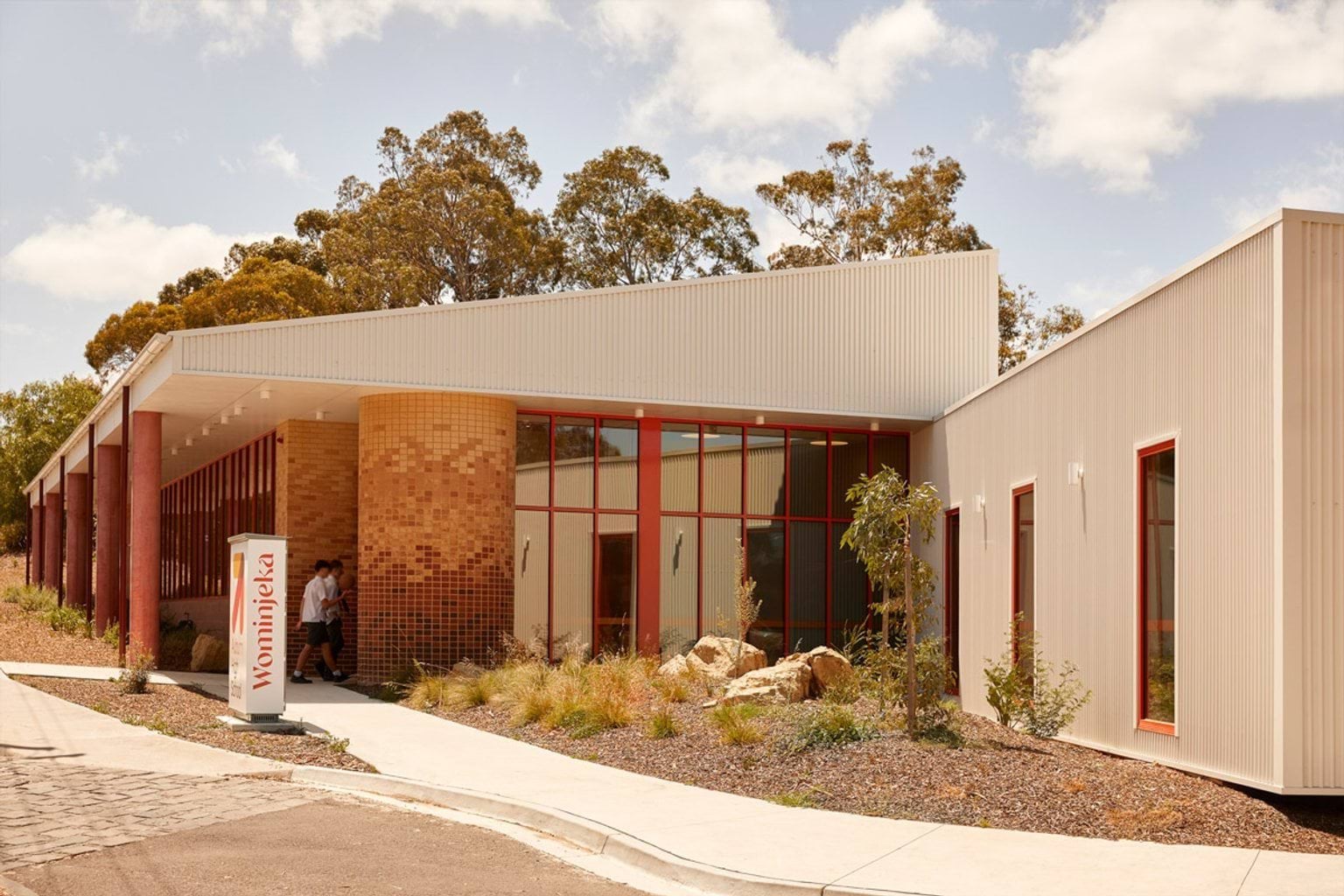 The main entrance of a school building with angled rooflines, brick columns, and red-framed windows. A landscaped garden with rocks and shrubs borders the walkway.