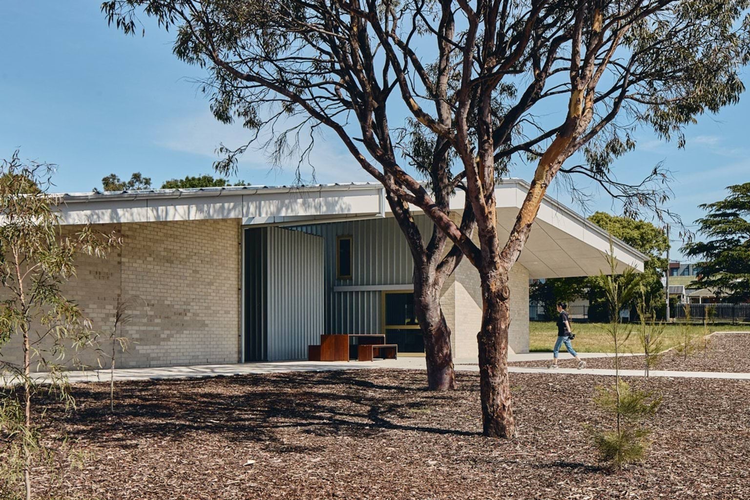 The exterior of a school building with pale brick walls and a wide roof, framed by tall gum trees and landscaped garden beds.
