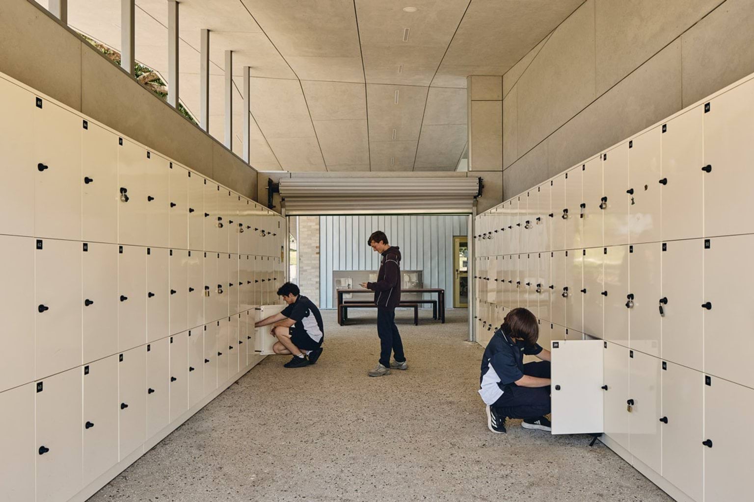 An open locker bay with rows of cream lockers on both sides and three students accessing their lockers under a high ceiling.