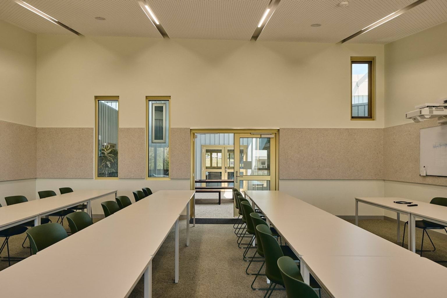 A classroom with white tables and green chairs arranged in a U-shape, looking out through glass doors to a covered courtyard.