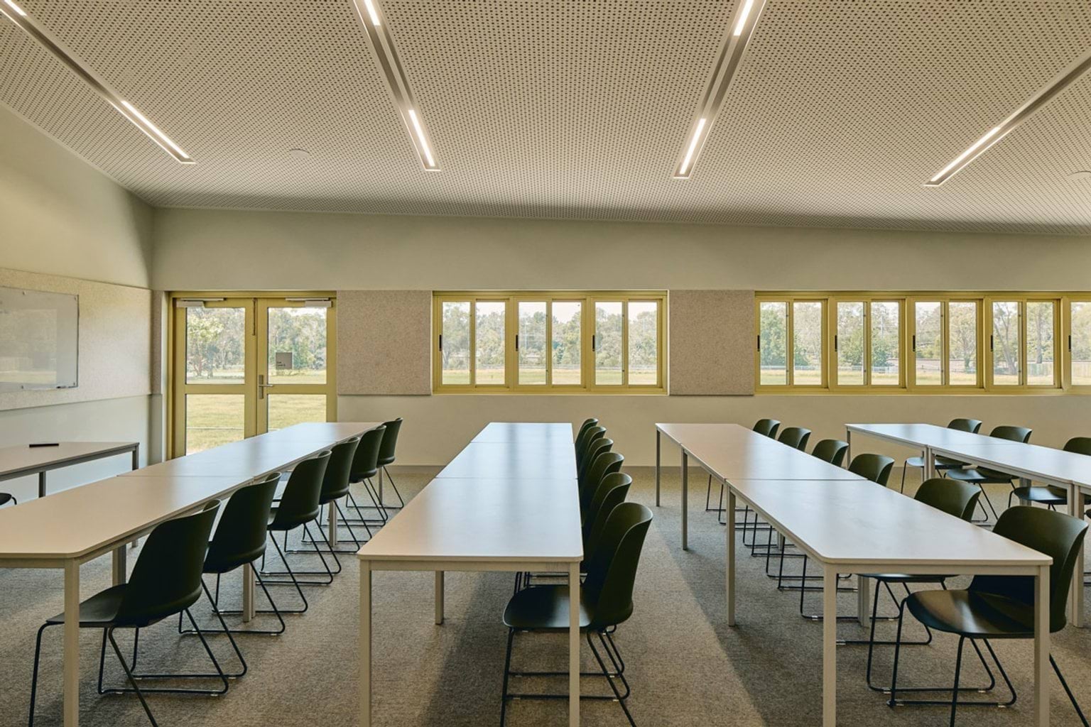 A classroom with long white tables and green chairs arranged in rows, facing a whiteboard and large windows overlooking greenery.