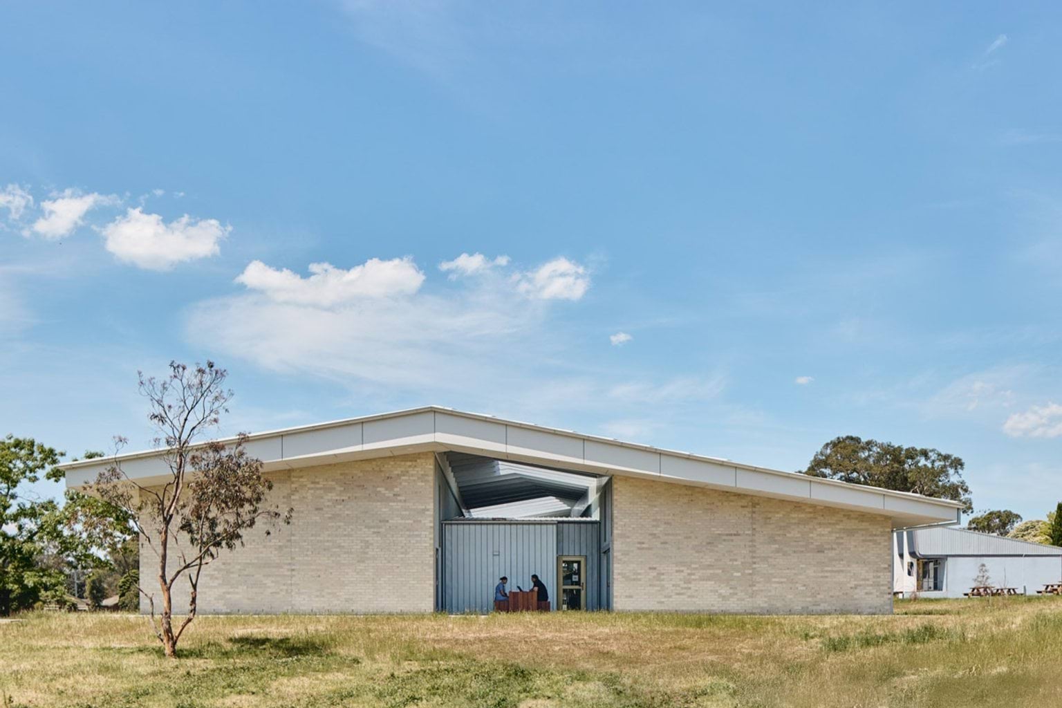 A modern school building with pale brick walls and a wide roof, seen from a grassed area with scattered trees.