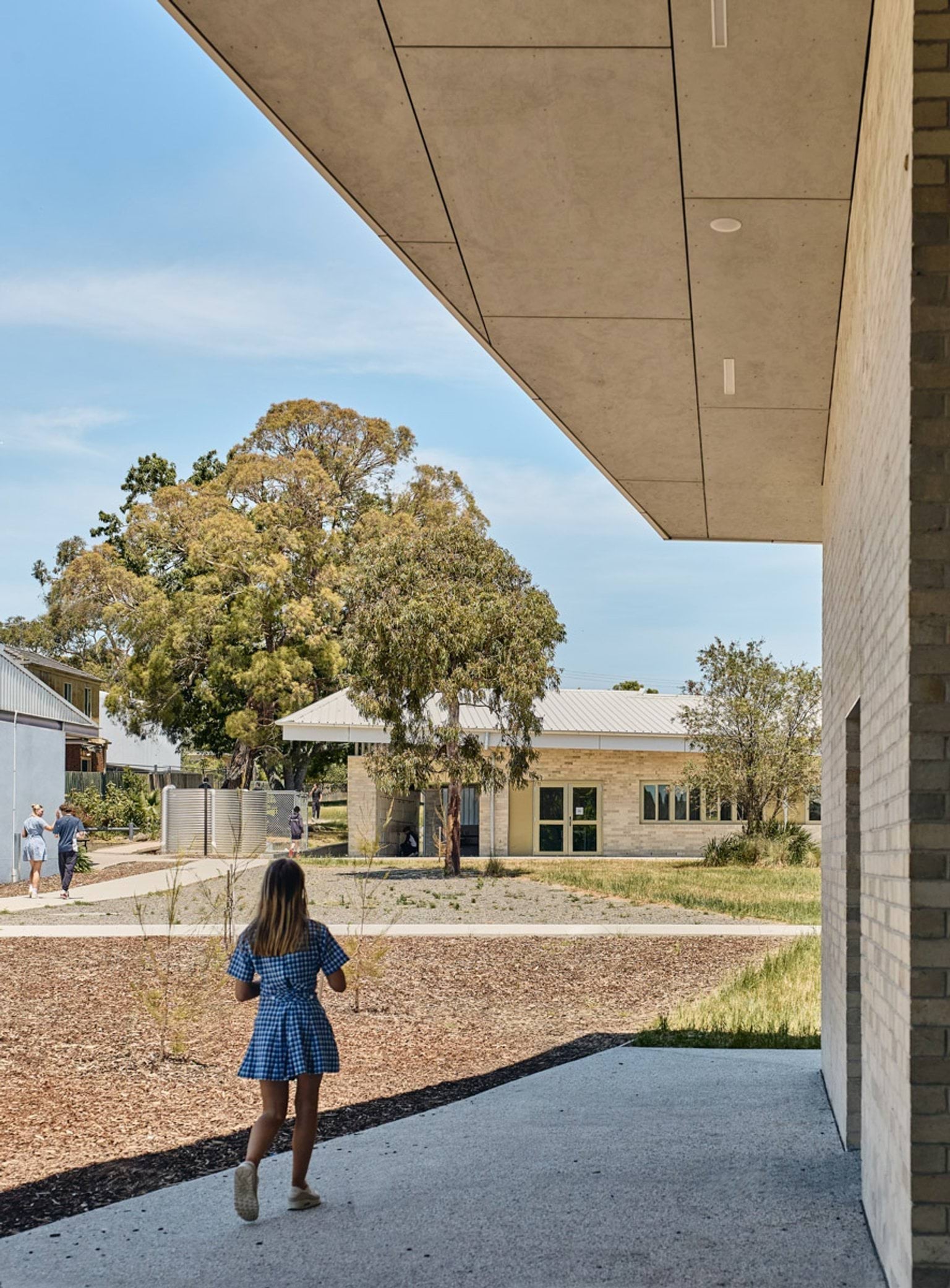 A student walking along a concrete path in a landscaped courtyard between modern school buildings with wide roofs and light brick walls.