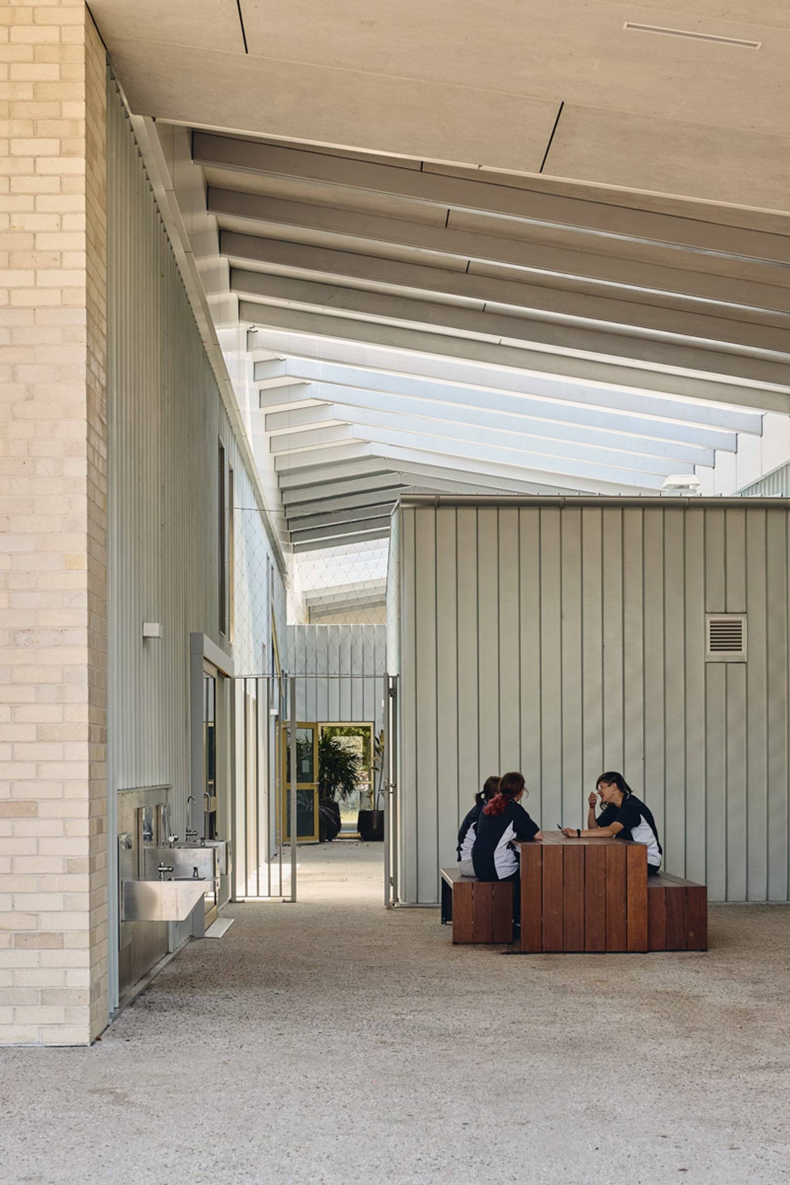 Three students seated at a timber table in a covered outdoor area with corrugated walls and slatted roofing.