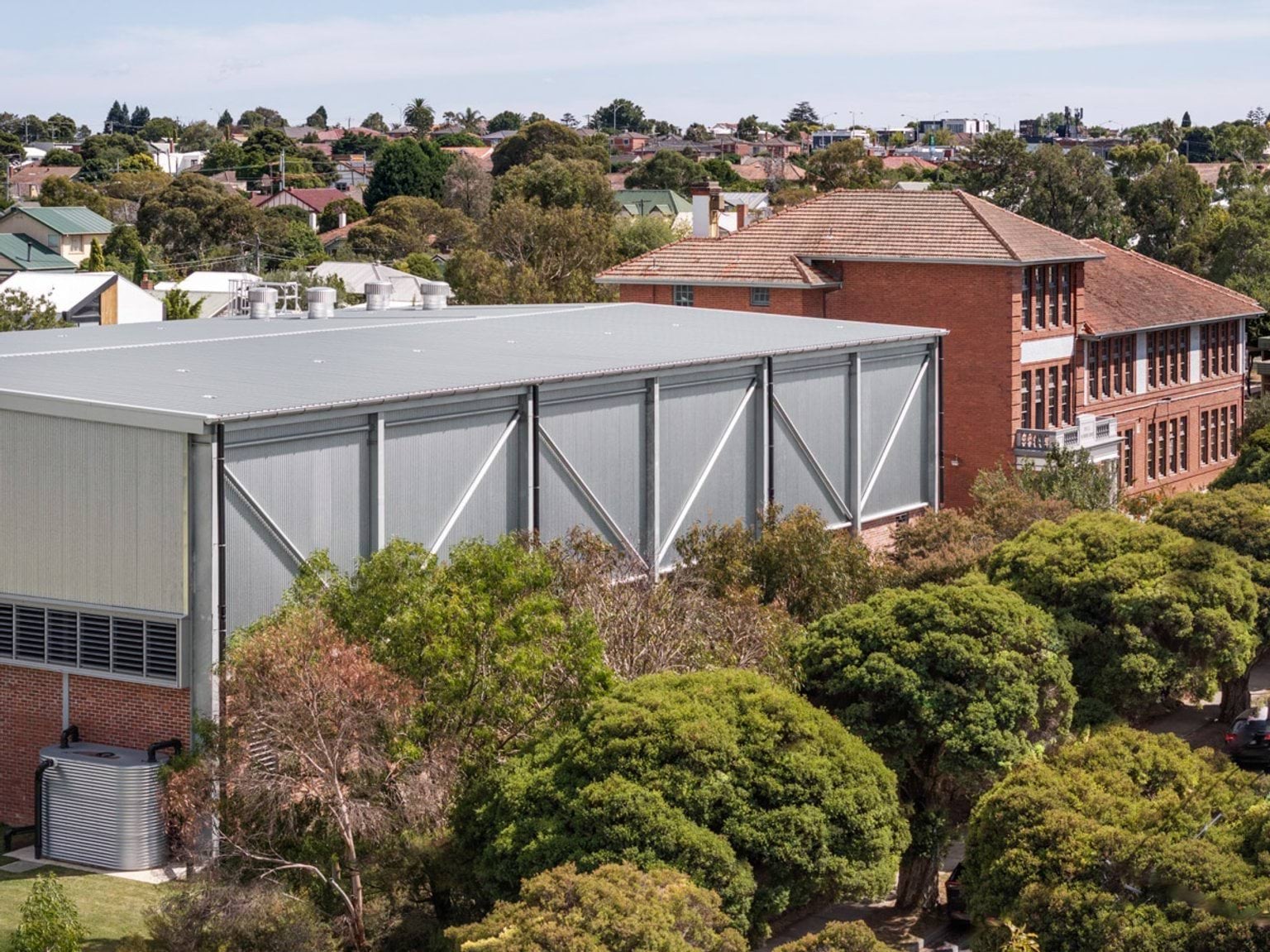 Aerial view of a school with a modern metal-clad sports hall next to a red brick heritage building, surrounded by trees and suburban houses.