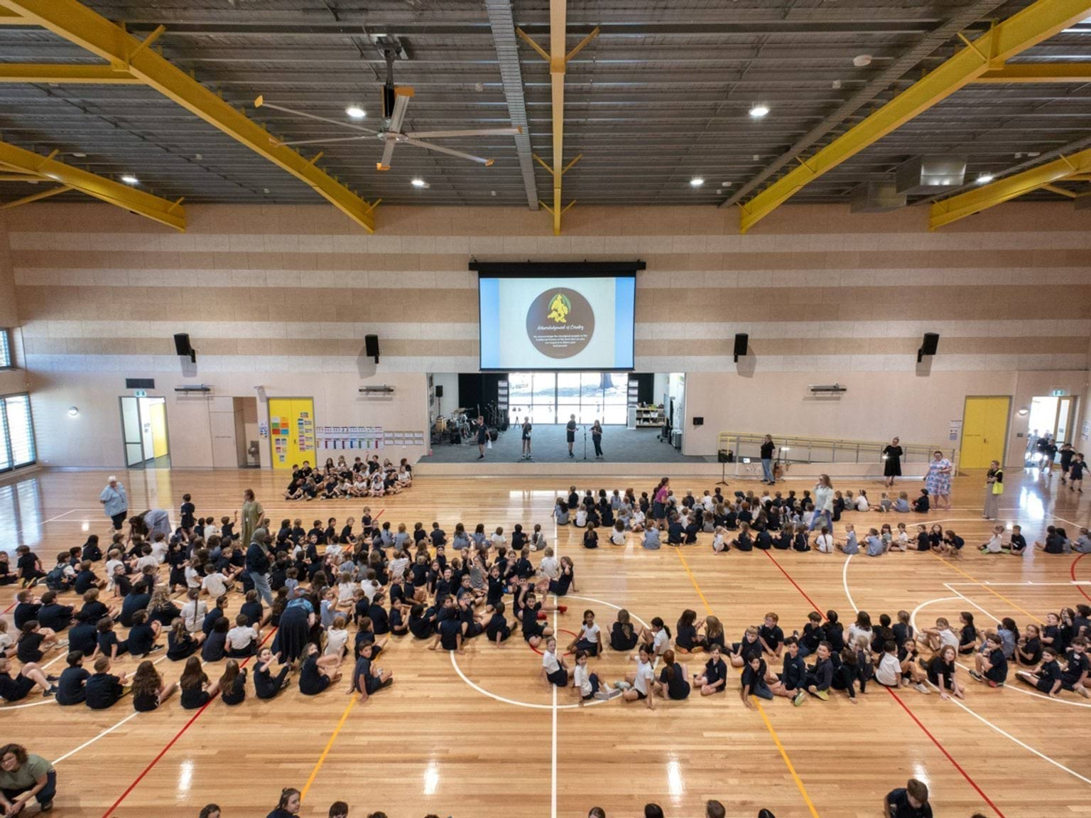 Students seated on a polished timber floor in a large gymnasium with yellow steel beams and a high ceiling. A projector screen displays a school crest at the front.