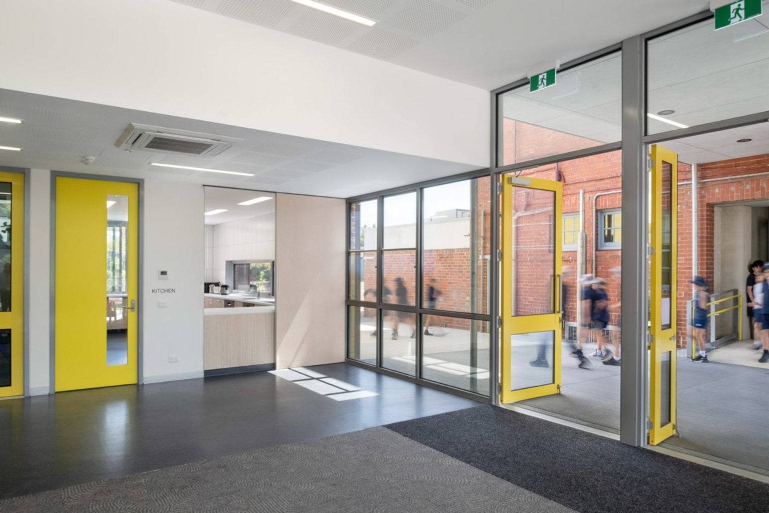A bright school foyer with glass walls and yellow-framed doors leading to an outdoor area. A kitchen space is visible through an internal doorway.