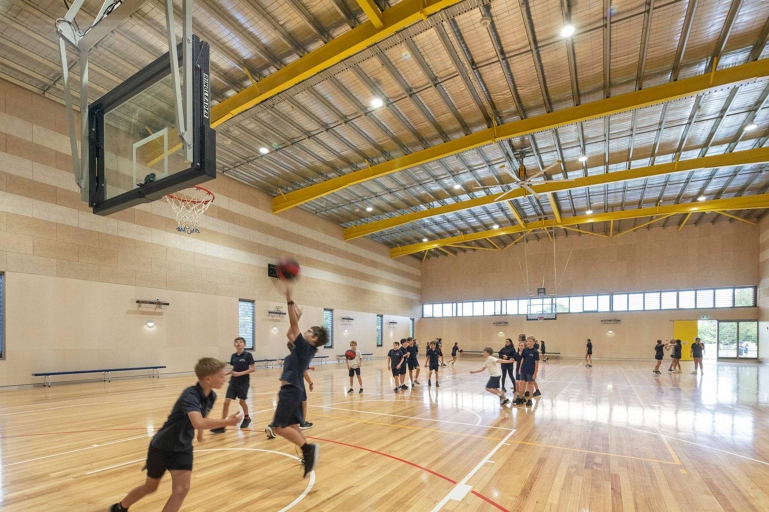 Students playing basketball in a large gymnasium with timber flooring, yellow steel beams, and high ceilings with natural light panels.