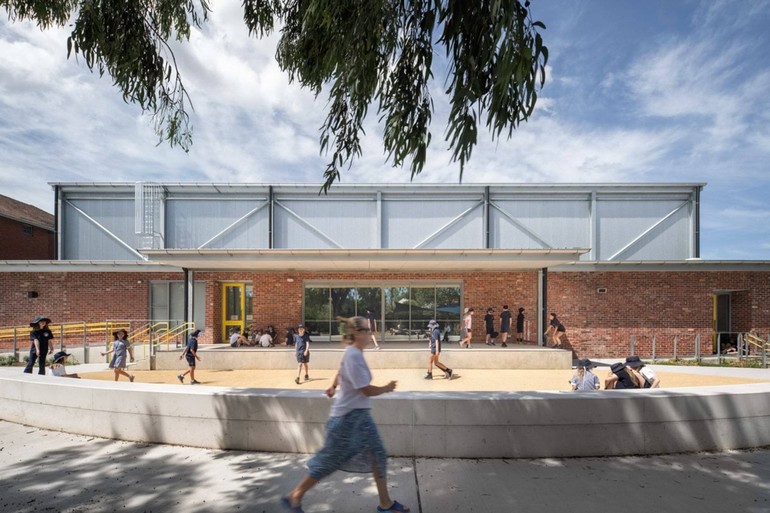 Students walking and sitting in a paved courtyard outside a school building with red brick walls, metal cladding, and yellow-framed doors.