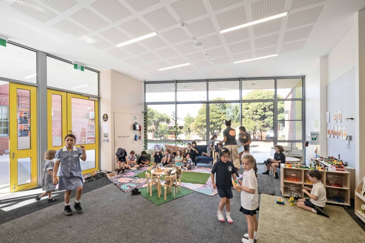 An indoor learning space with large windows, yellow-framed doors, and groups of students playing on mats and using toys and craft materials.