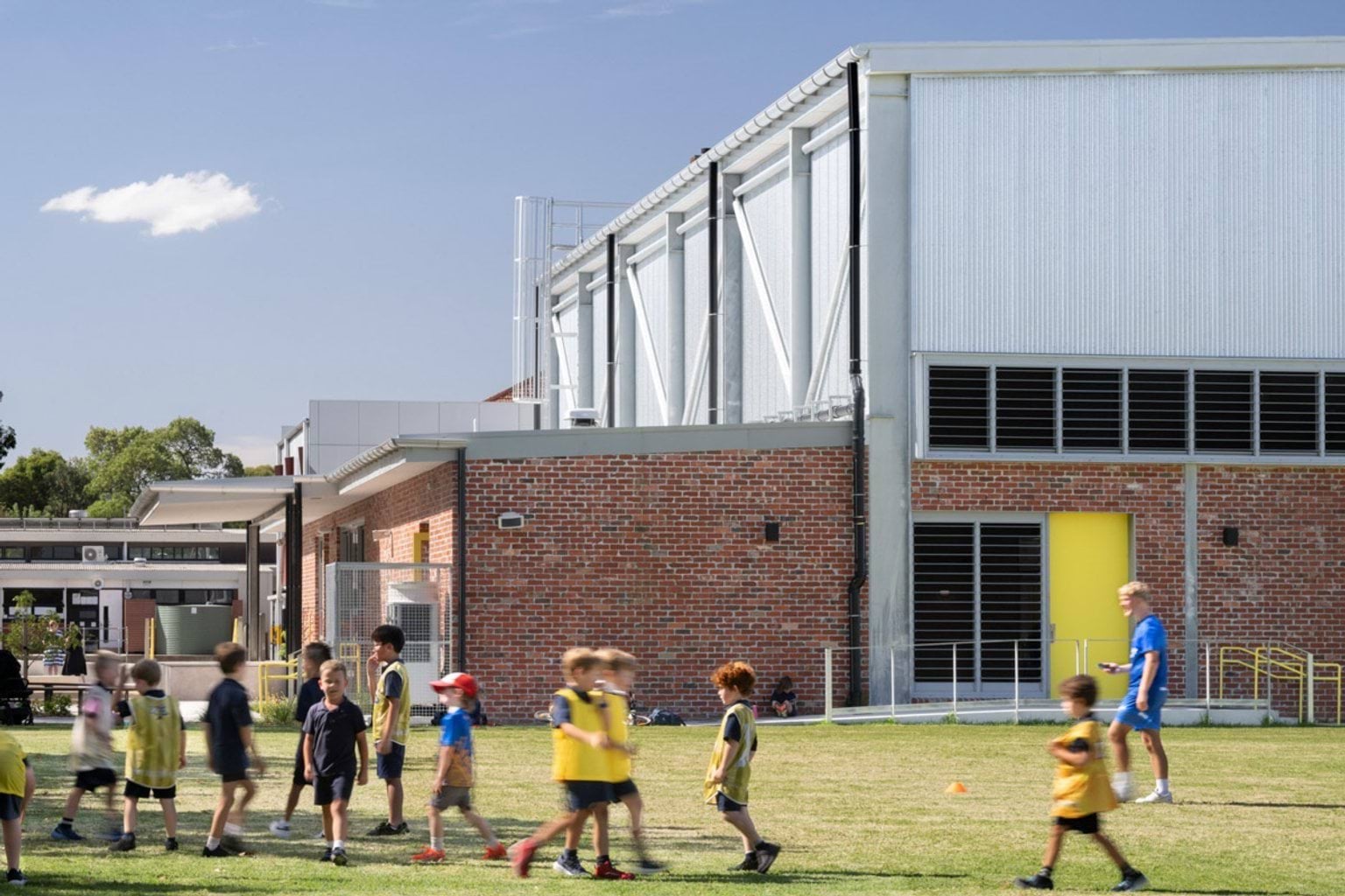 Students walking and playing on a grass sports field beside a modern school building with red brick walls, metal cladding, and a bright yellow door.