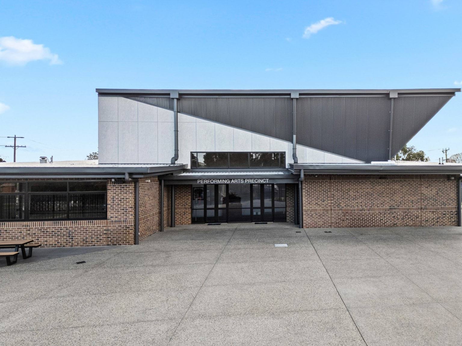 Front view of the Performing Arts Precinct with a wide entrance, brick walls, and a striking angular roofline against a clear blue sky.