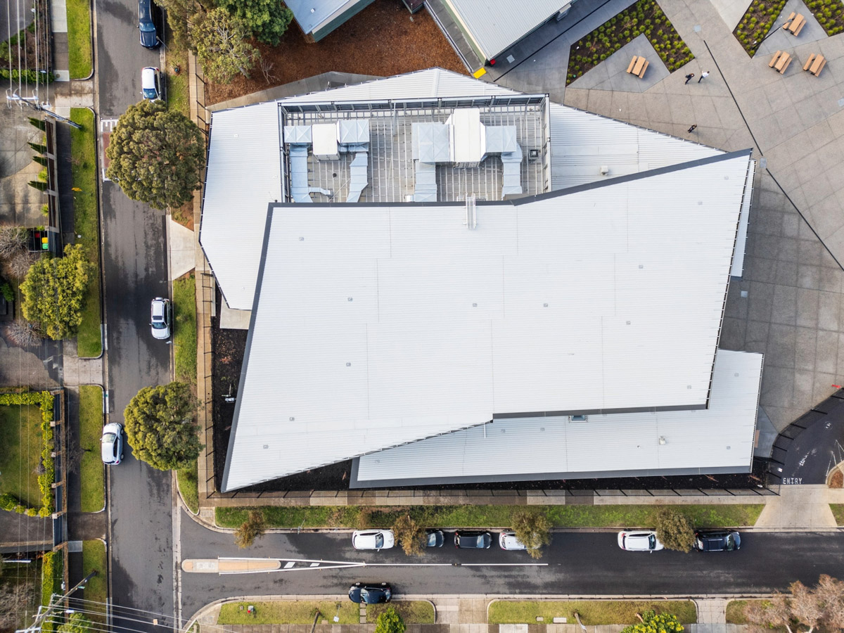 Aerial view of a modern school building with an angular roof design and light grey cladding, surrounded by streets, trees, and landscaped outdoor areas with seating.