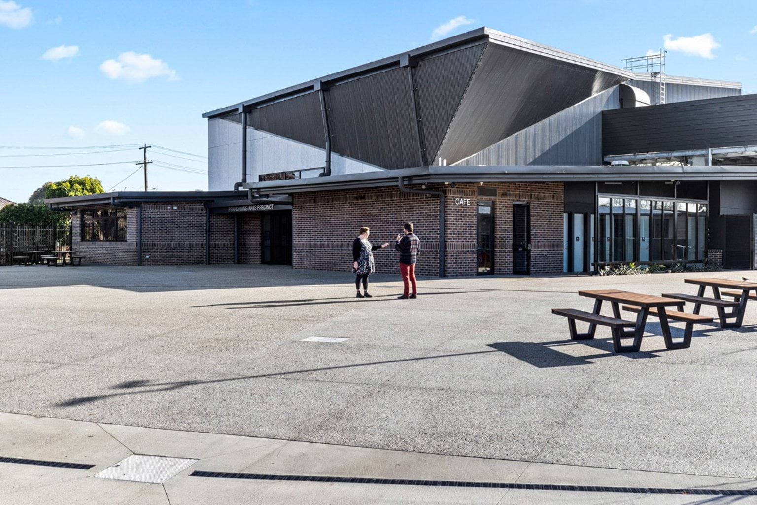 Open courtyard with timber picnic tables in front of a modern school building featuring brick walls and a dramatic angular roof.