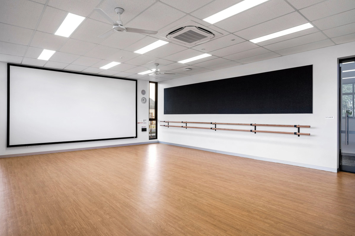 Dance studio with timber flooring, wall-mounted ballet barres, a large mirror, and a projection screen under bright ceiling lights.